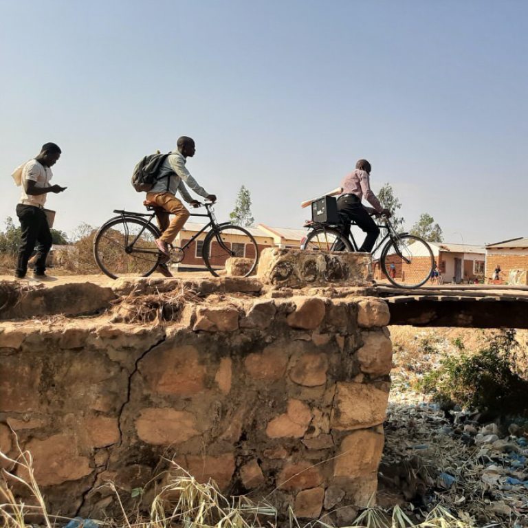African men riding bicycles over a rural bridge